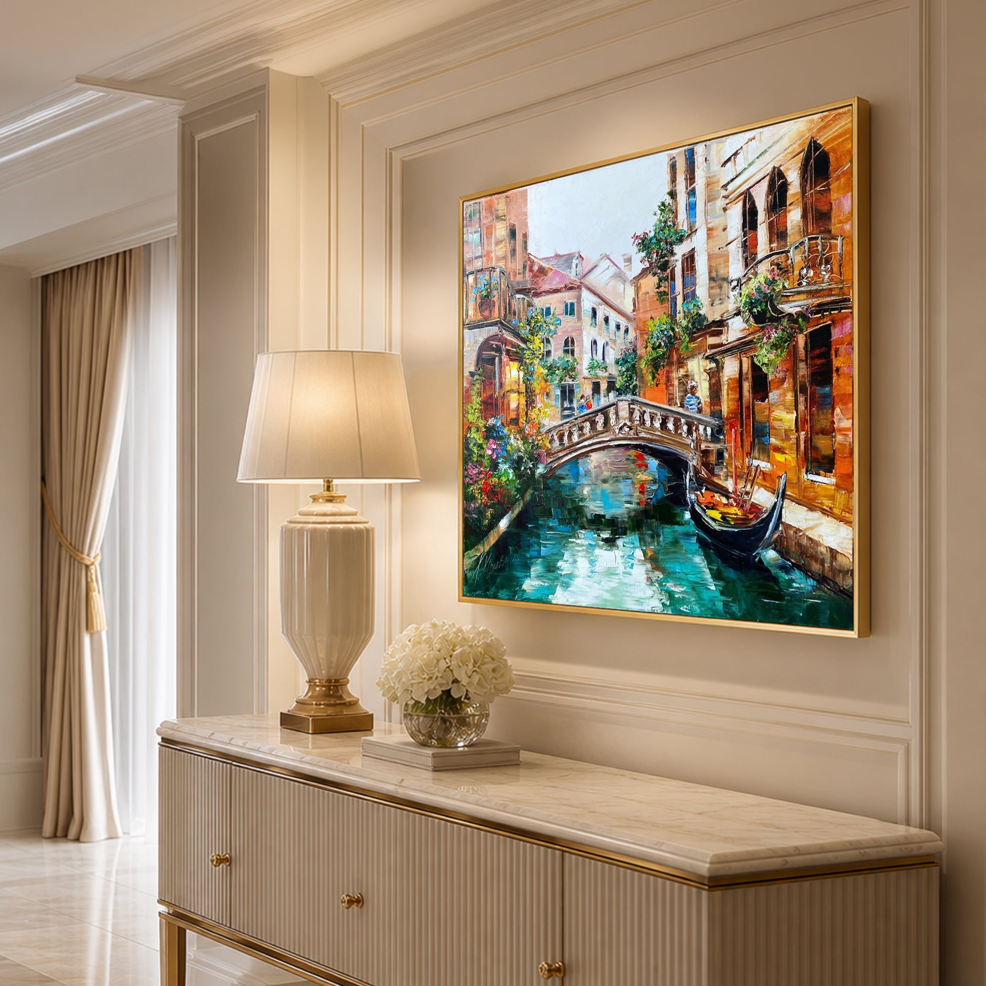 A framed canvas painting of a Venetian canal scene hangs on a beige wall above a console table with a lamp and flowers. The artwork features warm-toned buildings, a bridge, and a gondola in a colorful, textured style.