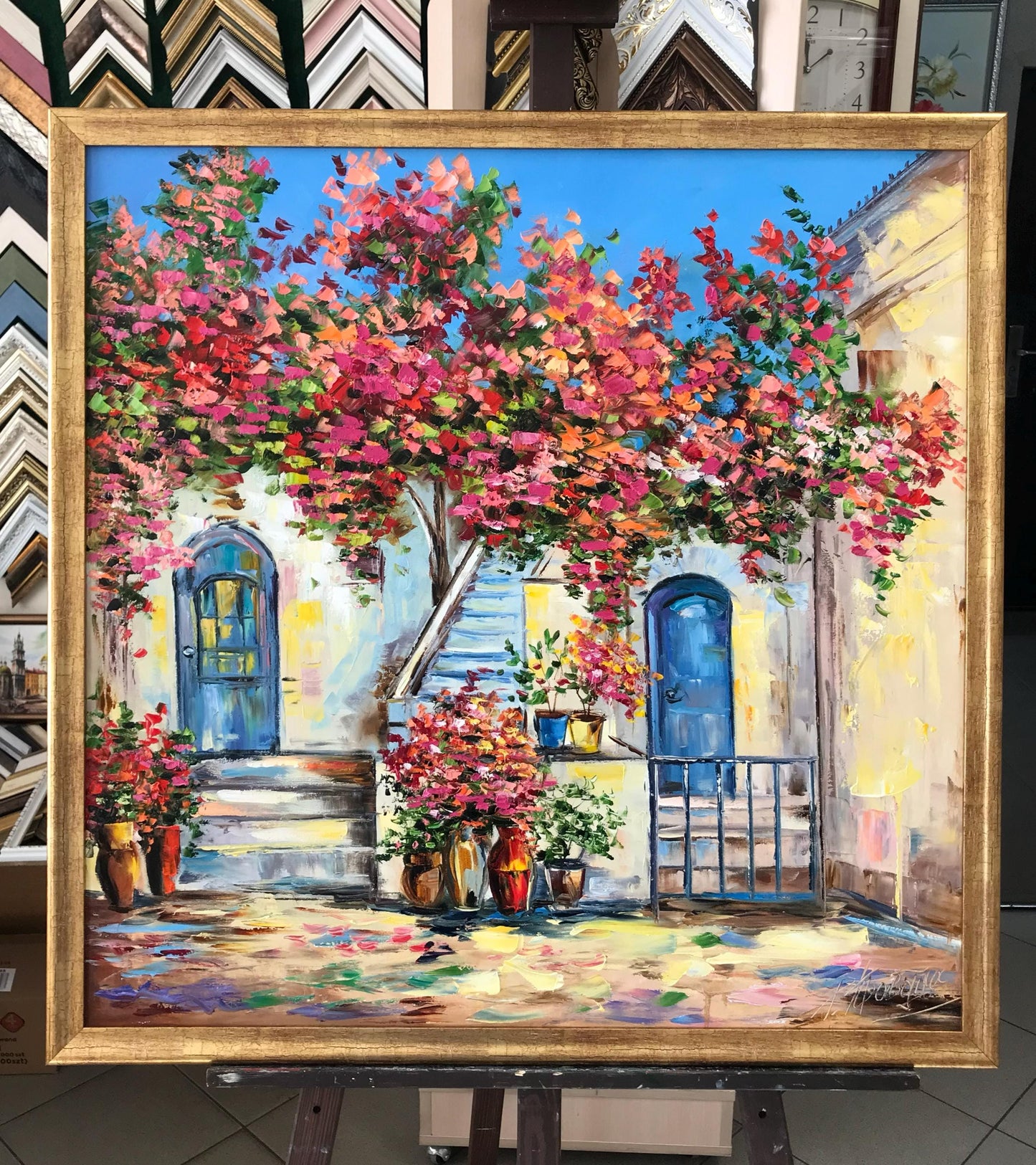 A framed oil painting of a Mediterranean courtyard with a vibrant bougainvillea tree in pink and red flowers, blue doors, and potted plants, displayed on an easel.