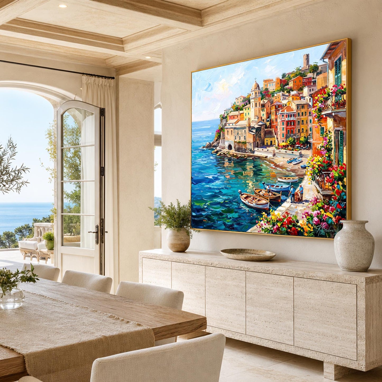 A large framed canvas painting of a colorful coastal village and harbor hangs on a beige wall in a bright, modern dining room with a stone credenza, dining table, and potted plants.