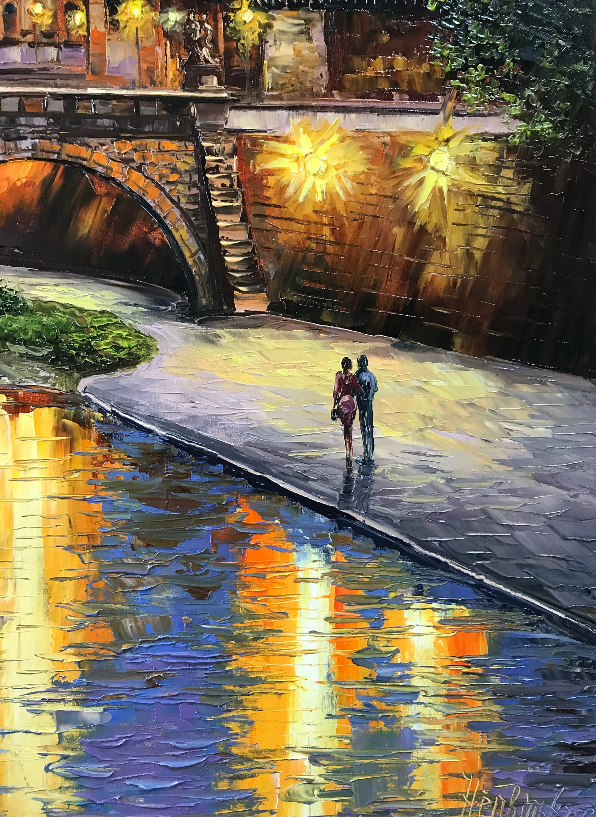 Two people walking along a stone path by a river at night, with warm lights and reflections.