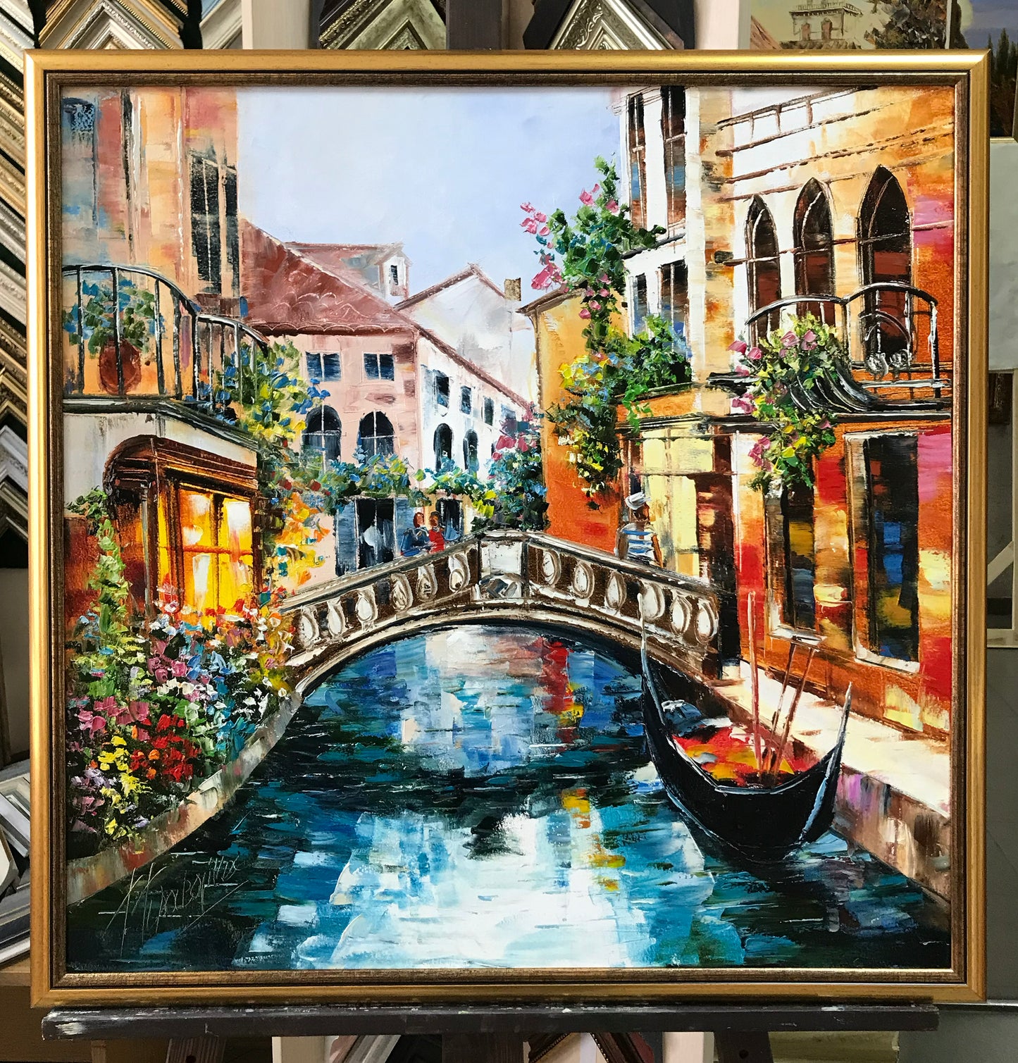 A framed oil painting of a Venetian canal scene with a gondola, colorful buildings, flowers, and a stone bridge, shown in a gold frame on an easel.