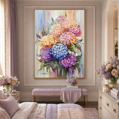 A large framed floral oil painting of colorful hydrangeas hangs on a beige wall above a lavender upholstered bench in a styled bedroom. The artwork features pink, purple, blue, and peach blossoms with green leaves, and the room is decorated with matching floral arrangements and soft bedding.