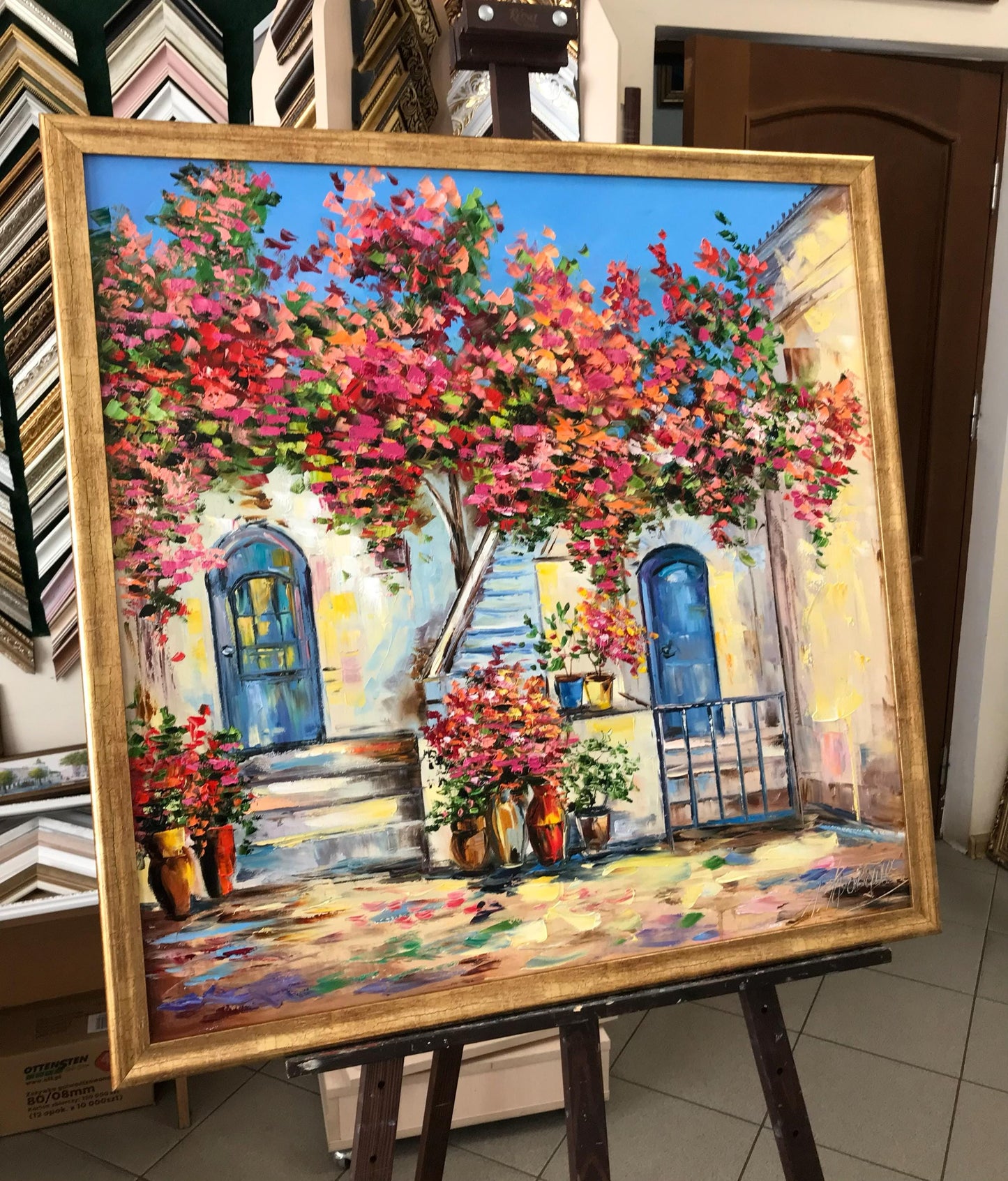 A large framed oil painting of a Mediterranean-style Greek courtyard with a vibrant pink and red flowering tree, blue doors, and a gold frame, displayed on an easel in a showroom. 