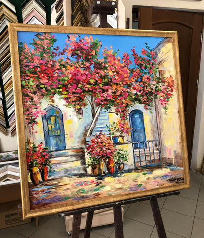 A large framed oil painting of a Mediterranean-style Greek courtyard with a vibrant pink and red flowering tree, blue doors, and a gold frame, displayed on an easel in a showroom. 
